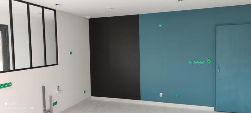 Une pièce moderne avec un mur blanc, un mur d'accent noir et un mur d'accent sarcelle avec une porte sarcelle assortie. Des panneaux de verre encadrés en noir sont sur la gauche, et diverses prises sont marquées de ruban adhésif vert.