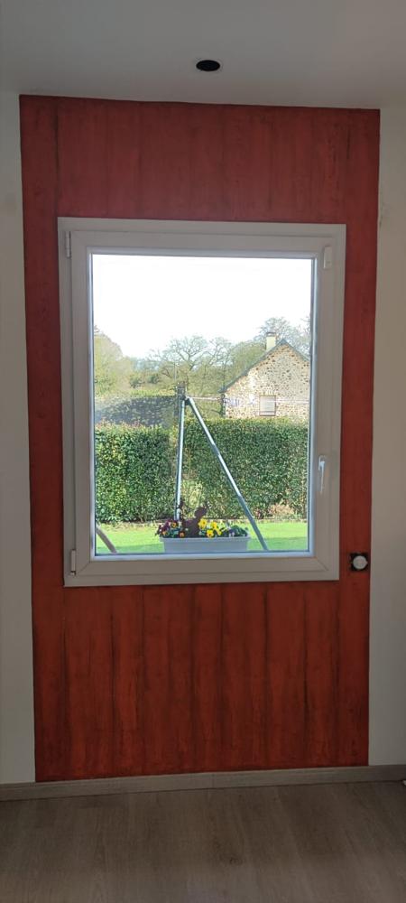 Vue d'ensemble d'un pan de mur peint avec un effet bois texturé rouge entourant une fenêtre blanche donnant sur un jardin.