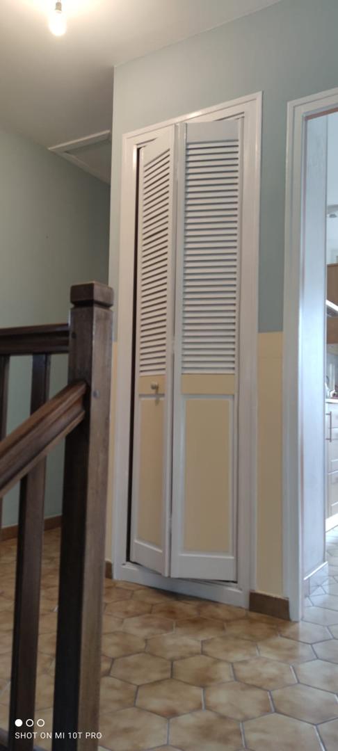 Une vue d'une porte pliante blanche à persiennes avec un panneau inférieur jaune, placée contre un mur bicolore peint en bleu clair et jaune. Un escalier en bois avec une rampe marron est visible au premier plan.