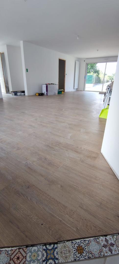 Une pièce fraîchement peinte avec un sol aspect bois clair posé en diagonale, une petite section de carreaux décoratifs au bas et des murs blancs et lumineux avec de grandes portes vitrées à l'extrémité.