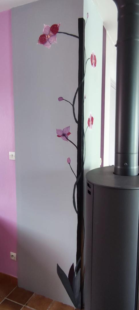 Un gros plan d'un mur avec une palette de couleurs rose et violet. Le coin du mur est peint avec une vigne noire et des fleurs d'orchidées roses. Un poêle à bois cylindrique noir est visible à côté du mur.