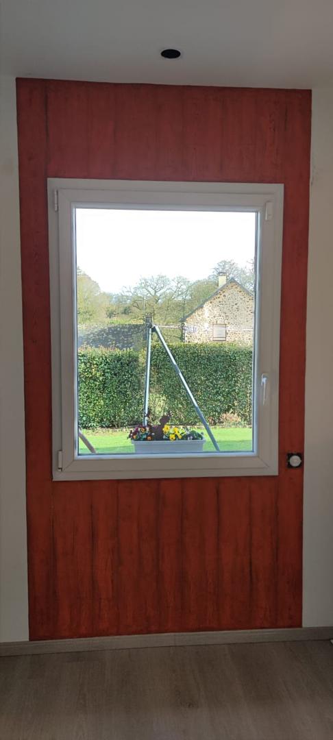 Vue d'ensemble d'un pan de mur peint avec un effet bois texturé rouge entourant une fenêtre blanche donnant sur un jardin.