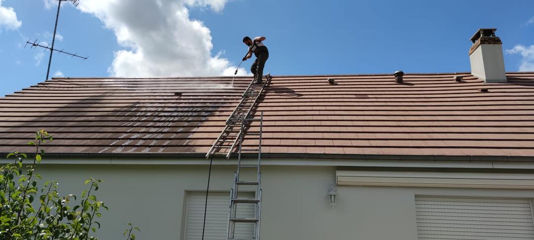 Artisan sur une échelle nettoyant une toiture en tuiles au nettoyeur haute pression.