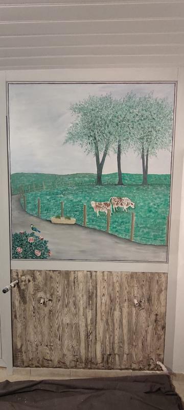 Une grande peinture murale d'une scène de champ avec deux vaches, des arbres verts et une route, peinte sur la moitié supérieure d'une porte ou d'une armoire. La moitié inférieure est peinte pour ressembler à une surface en bois rustique.